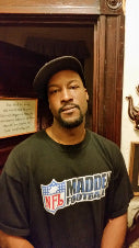 A man wearing a black t-shirt with a large 'Madden Football' and NFL logo on the front, photographed in a dimly lit indoor setting.