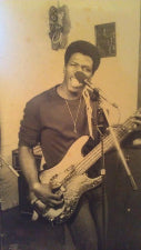 A sepia-toned vintage photograph of a man playing a bass guitar and singing into a microphone in an indoor setting.